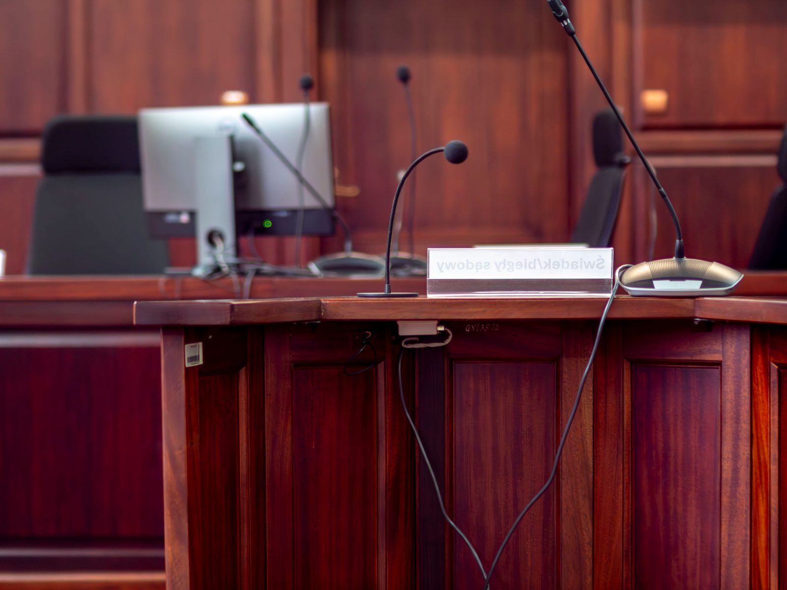 An empty courtroom witness stand with gooseneck microphones and a monitor